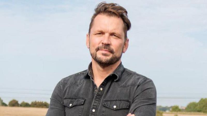 A man stands in a field on a cloudy day. He has his arms folded in front of him. He has brown hair that is swept to the right and he is wearing a faded black denim shirt. He has a brown beard.