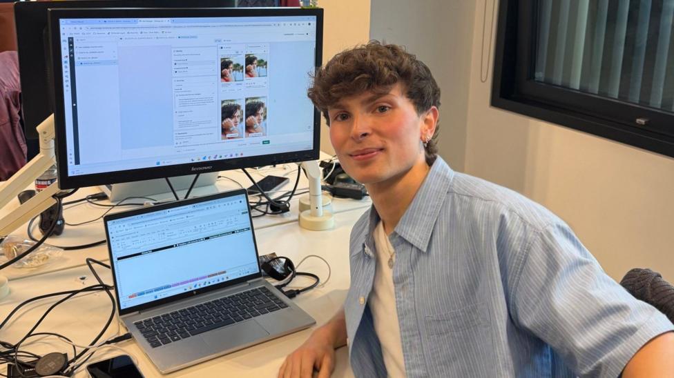 Oliver is looking at the camera smiling. He is sat at his work desk.