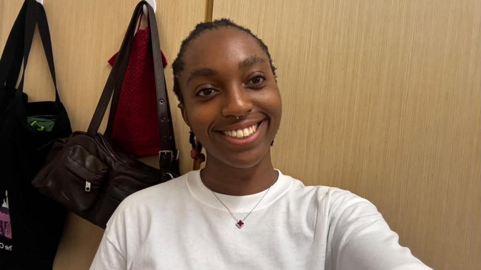 Chinaza Eke is facing the camera smiling. She is wearing a white long sleeved top, with a small cross on a necklace. Bags are hanging on the wall behind her.