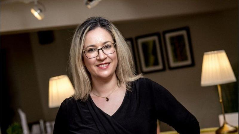 Selina Horshi, a woman with blonde hair, who has glasses, she is wearing a dark top. There are two lamps and pictures in the background.