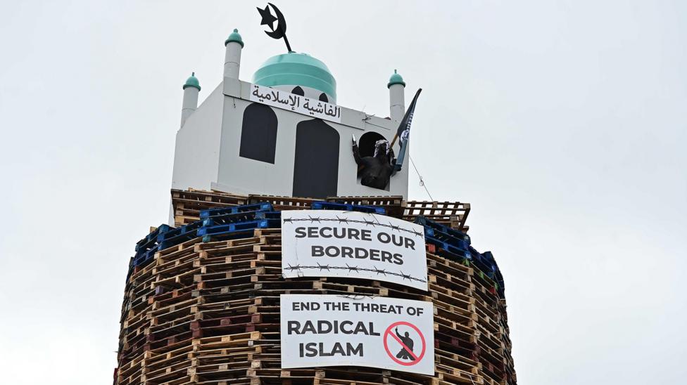 An image of the top of the bonfire, consisting of wood pallets, with a structure resembling a mosque on top. Two placards are below. One reads "secure our borders" while another reads "end the threat of radical Islam". A figure of a man is coming out from one of the windows. The figure is wearing a headscarf and is holding a model of a knife and a flag.