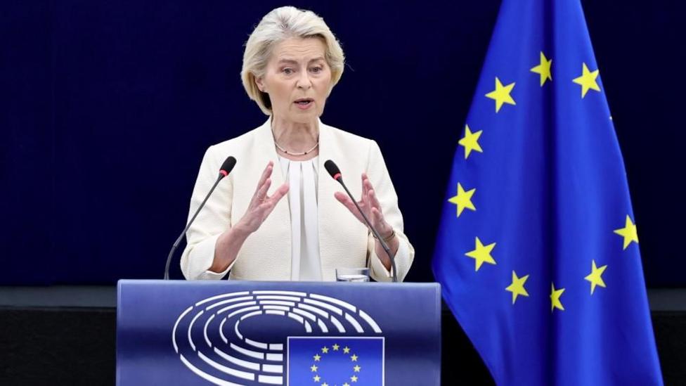 Ursula von der Leyen, who has a grey/blonde short haircut, wears a cream blazer and blouse as she speaks at a podium, with the EU flag in the background