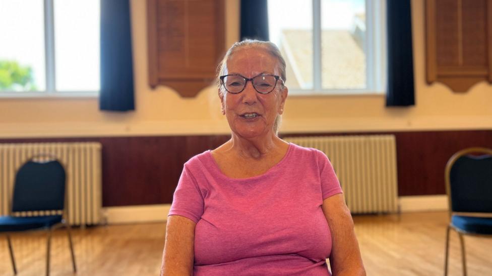 Linda Crawford is wearing a bright pink polo shirt. She sits in a community hall, facing the camera. Exercise equipment, cushions and chairs are arranged behind her, with a stage and stained-glass window visible in the background.