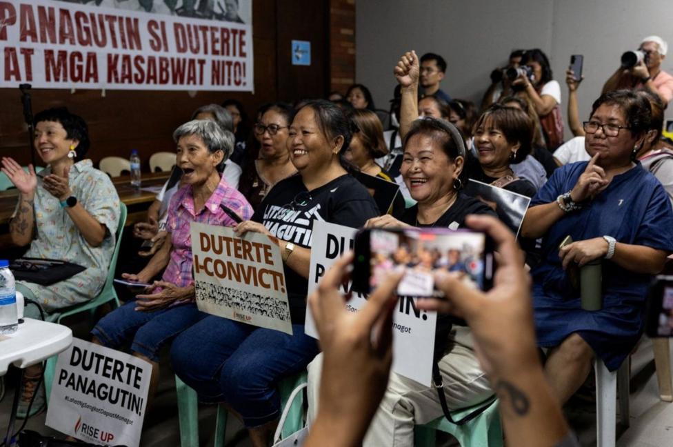 More than 10 people dressed casually sitting in a room holding anti-Duterte placards, while cheering.