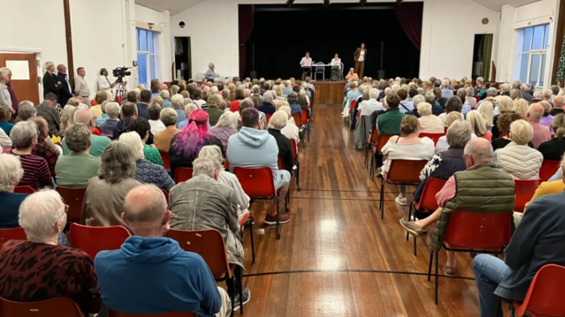 Picture taken from the back of a hall. Tens of people sat down facing a stage. Three people on the stage, two sitting at a table, and another standing up.