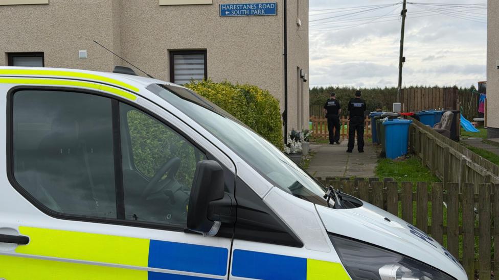 Two police officers outside a house in Armadale, West Lothian, with a police van parked in the foreground of the image.