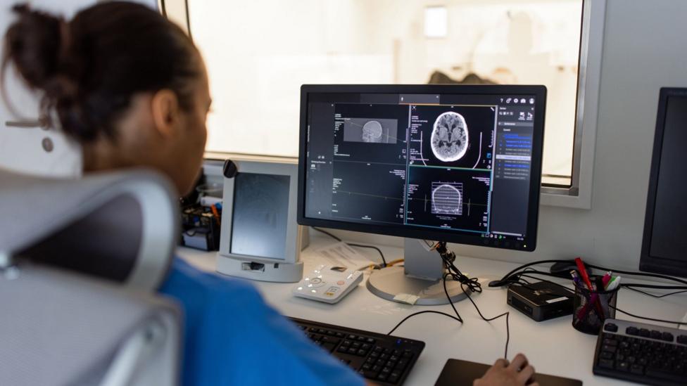 A nurse in a short sleeved blue top looks at a brain san on a computer, in the background is a glass window behind which the outline of an MRI scanner can be seen.