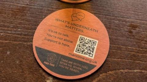An orange beer mat the words 'what's underneath matters' written at the top displays a QR code, phone numbers and a website link.