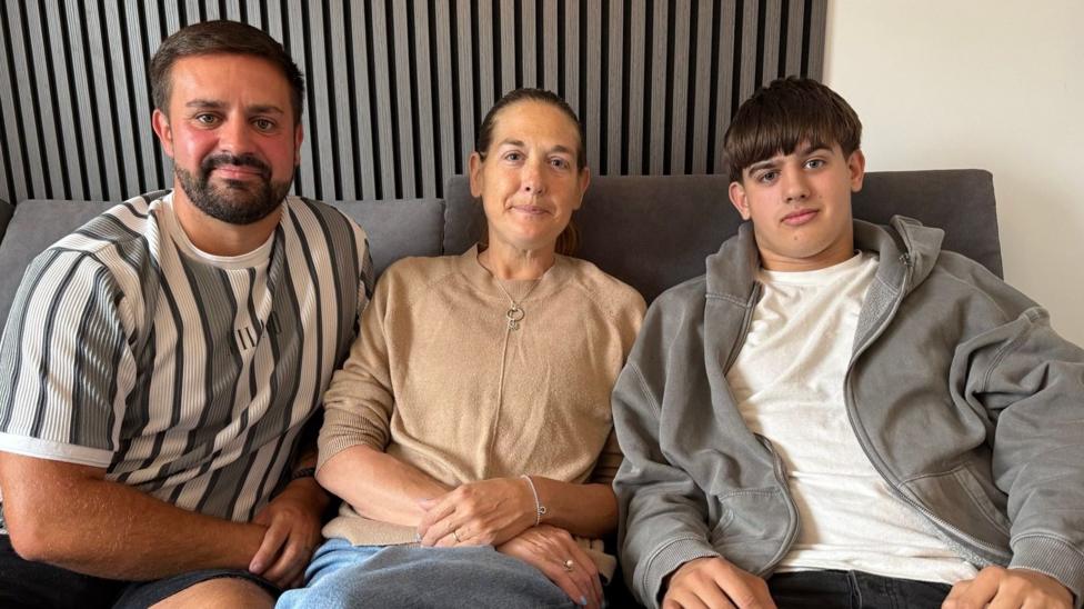 Karen's is pictured with her husband, Matthew, who has black hair and a beard and wears shorts and striped black and white t-shirt and her 17 year old son, Brandon, wears a grey hoody and has a mop of dark hair
