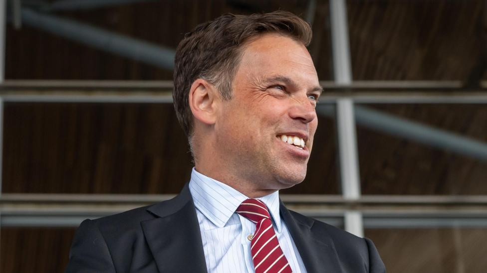 Ken Skates smiling off to the side of the image, wearing a dark suit and red and white tie, with a pin stripe shirt.