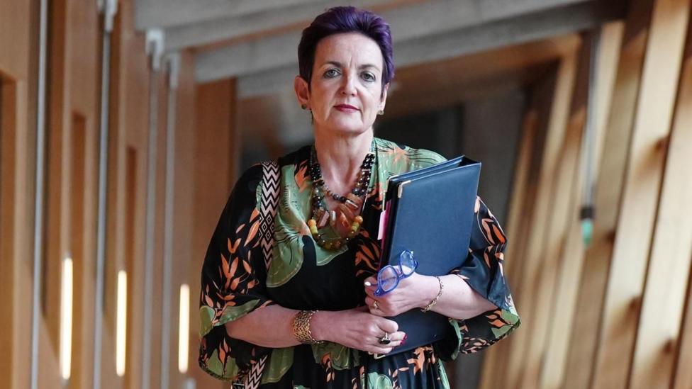Angela Constance walking through the Scottish Parliament. She has short purple hair and is wearing a patterned dress and carrying a blue folder.