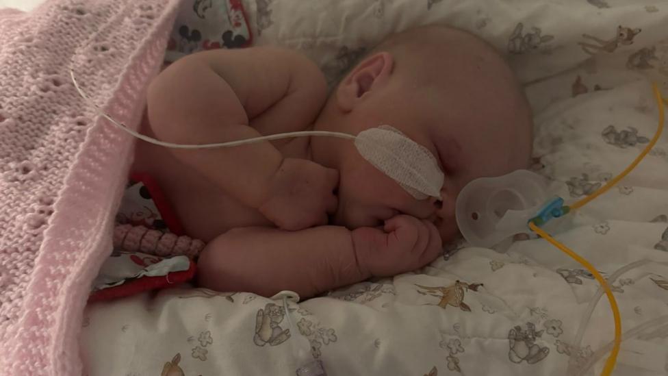 A small baby, Grace, is lying in a neonatal cot with tubes helping her to breathe and feed. She has a pink blanket over her.