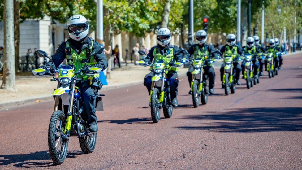 A line of Metropolitan Police officers riding high-powered e-motorbikes down a paved London road in single file formation, wearing full protective gear and high-visibility jackets.