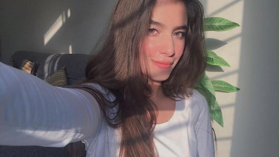 Person with long dark hair taking a selfie indoors, wearing a white long-sleeved top. Natural sunlight streams through a window, casting striped shadows across the room, with a sofa and leafy houseplant visible in the background.