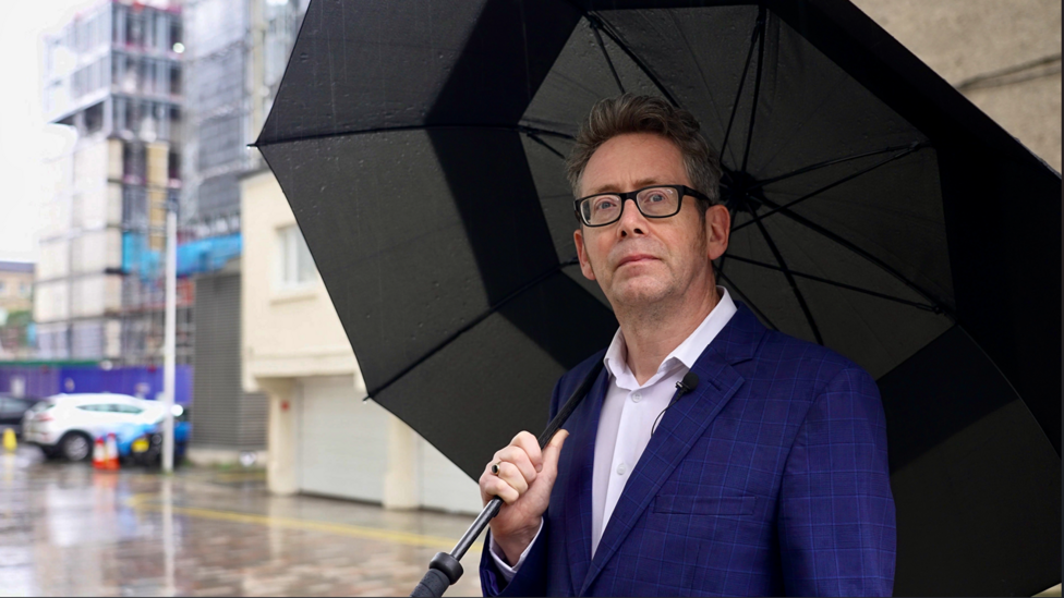 A man, Dr John Boyle, stands on a wet street looking thoughtful. He is wearing a dark blue suit and is holding a large black umbrella.