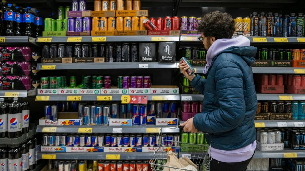 A male shopper in a jacket and hoodie looks at a selection of energy drinks stacked up in a fridge in a supermarket.