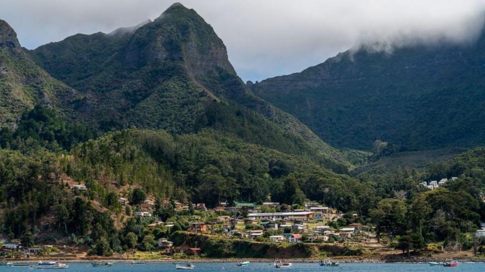 An island village nestled beneath tall mountains next to a bay with boats bobbing on the water
