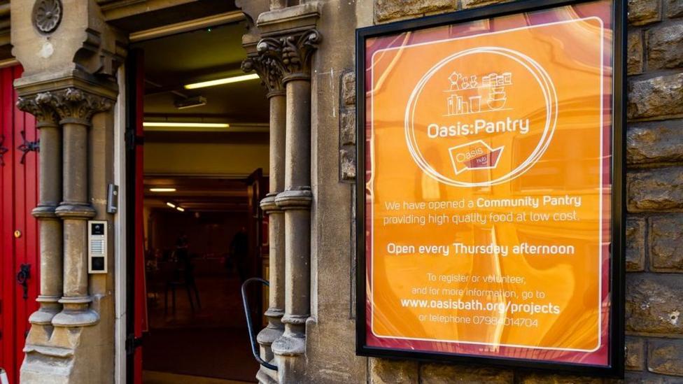 A rectangular orange sign with white text is on the outside wall of a building. The doorway is next to it and the doors are open.