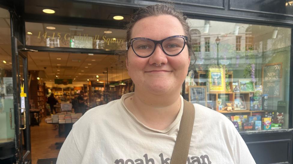 Georgia Rollinson stands in front of a Waterstones book shop in Carmarthen. She has brown hair, blue glasses and is wearing a white t-shirt.