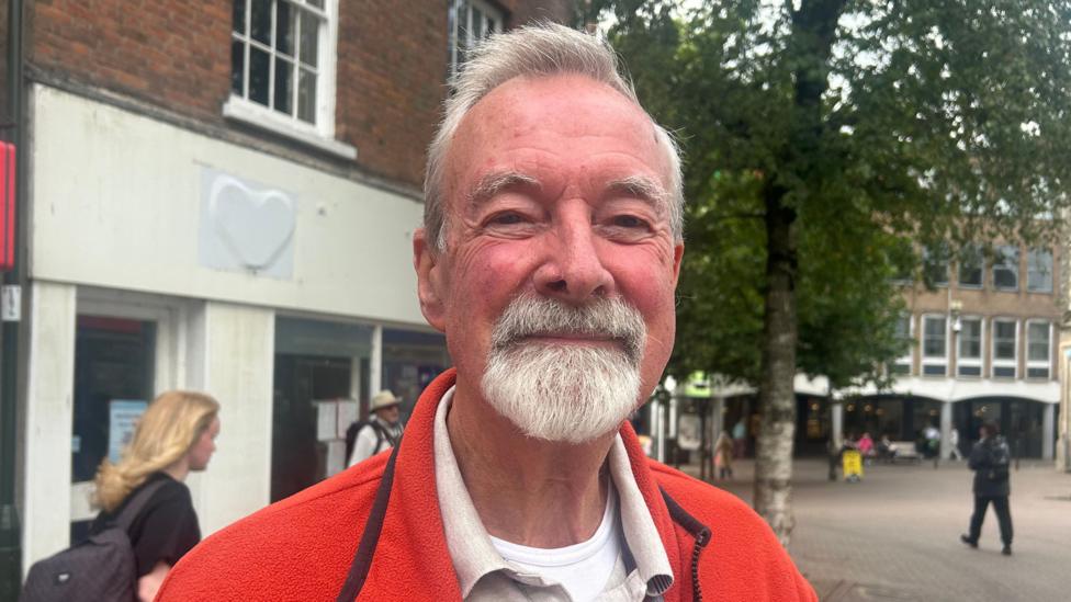 Phillip Richardson has white hair and a white beard. He is wearing a red fleece and a white polo shirt.