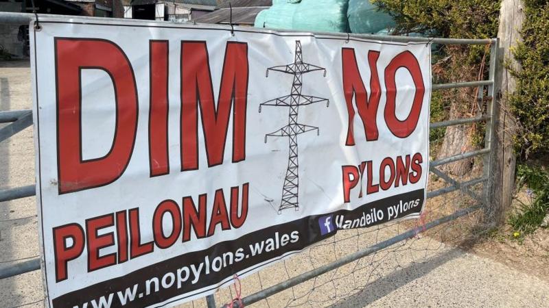 A sign on a metal gate which reads 'no pylons' with a pylon in the middle of it. 