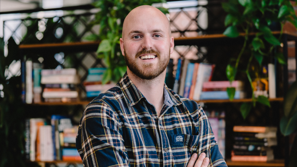 Guto Davies of Green Gen Cymru smiles at the camera, wearing a checked shirt and with his arms folded. He has a bald head., blue eyes and a brown beard and is standing in front of a bookcase which also has lots of green houseplants