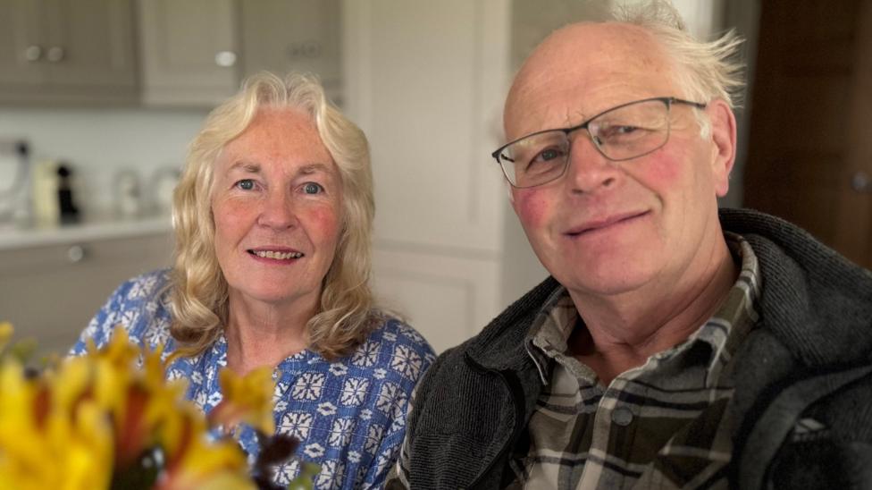 Ruth and Mervyn Kelso are sitting in a kitchen. There are yellow flowers in the bottom left hand corner. Ruth has shoulder length blonde/grey hair and is wearing a blue patterned shirt. Mervyn is wearing a brown checked shirt with a gilet. He has grey wispy hair and glasses.