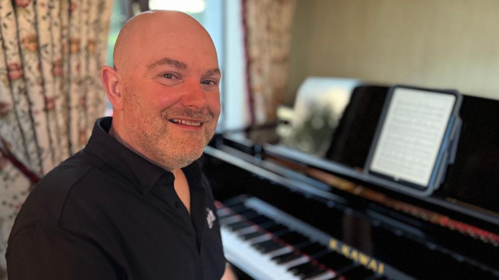 Barkley Thompson is sitting at a piano, turned towards the camera. There are patterned curtains behind him, tied back. He has no hair, a trimmed beard and is wearing a black polo shirt.