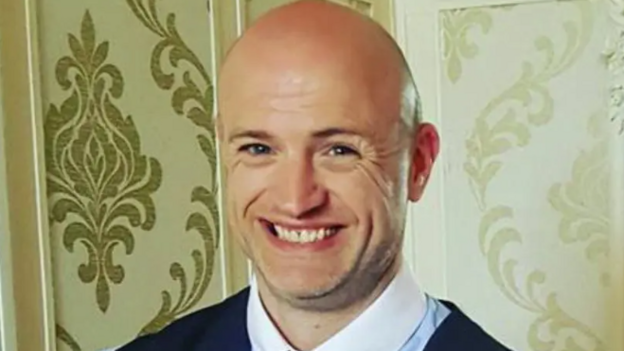 Shows a man with bald head smiling. He is wearing a blue shirt with white collar, blue tie and blue waistcoat. 