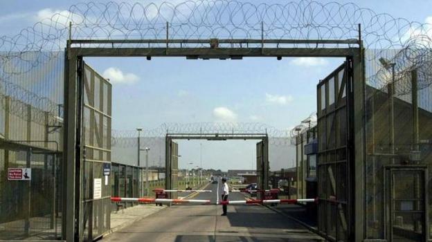 A prison guard in a white shirt and black trousers stands at a security gate in the inner area of the main entrance to Maghaberry prison where the main gates are open
