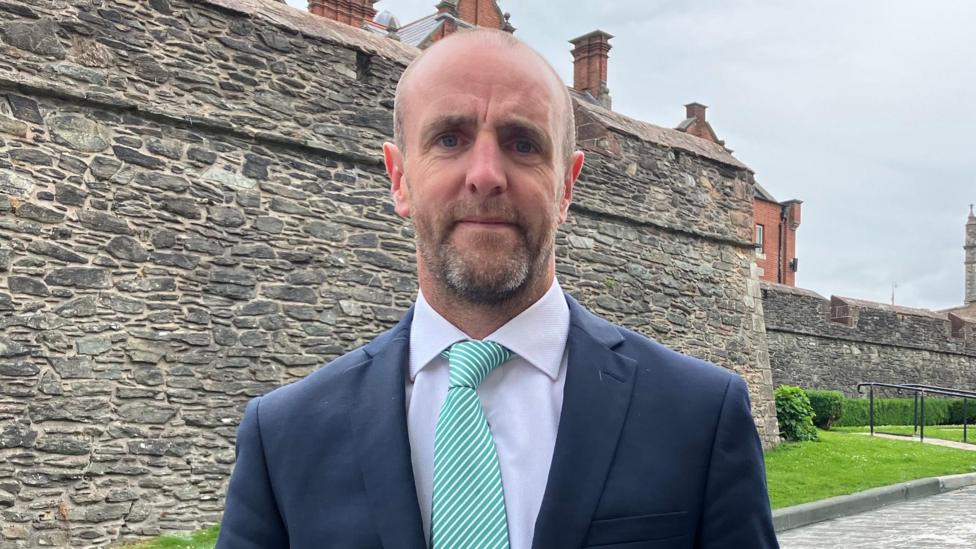 Mark H Durkan is wearing a suit and tie. His blazer is navy, shirt is white and his tie is green and white stripes. He has facial hair. Behind him are the walls of Derry and a red brick building. There is a paved grey path and green grass between him and the walls. In the distance we can see a large stone church tower.