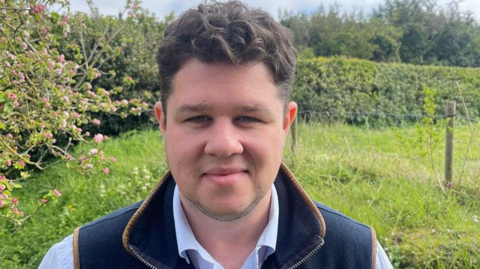 A man with dark hair smiles at the camera, wearing a dark blue gilet and a light blue shirt, in a field next to hedgerow