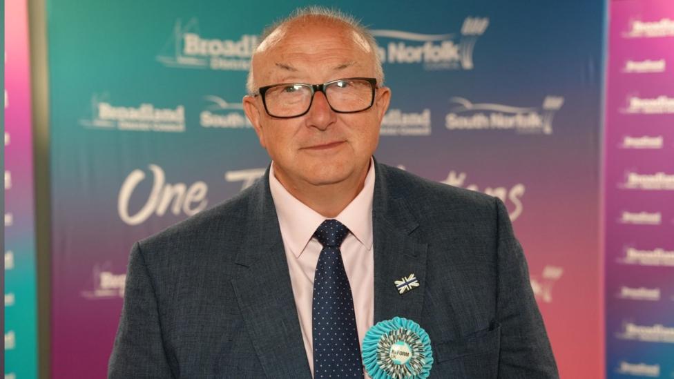 A man wearing glasses, a dark suit and a spotted tie poses in front of a colourful event backdrop. Visible branding includes “Broadland,” “South Norfolk,” and “Reform UK,” with a rosette and lapel pin attached to his jacket.