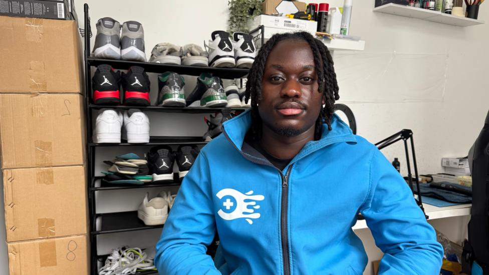 A man wearing a blue jacket with a white logo. In the background this a shoe rack with multiple trainers, a stack of boxes on the left and a desk with other belongings on the right.