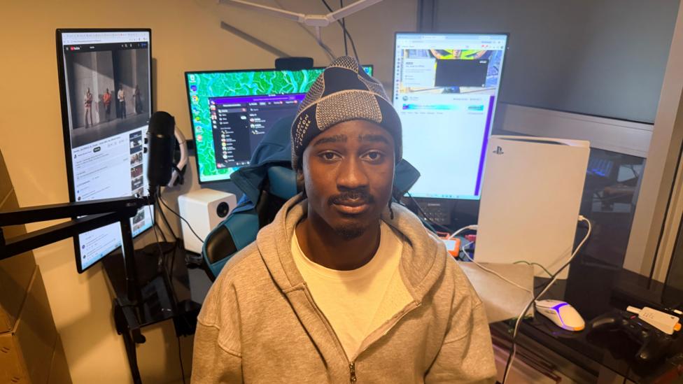 A man wearing a knitted cap and grey hoodie with a white t-shirt underneath. In the background there are multiple monitors and a PS5 console on the desk.