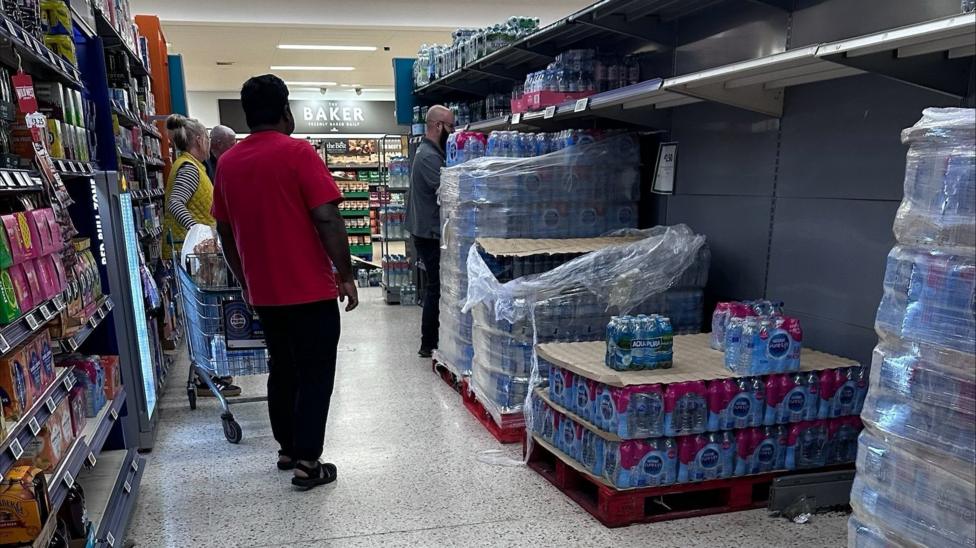 The photo shows a group of people in a supermarket aisle. They are looking at numerous bottles of water all stacked up on shelves and pallets.