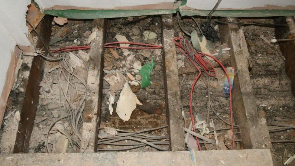 An exposed area of floor from where the boards have been removed. It is a cluttered mess with fragments of wood and plaster having fallen into the whole between the wooden bars supporting the floor. There is also red wiring, but amid the mess is a section where there is a clear patch where presumably the baby was