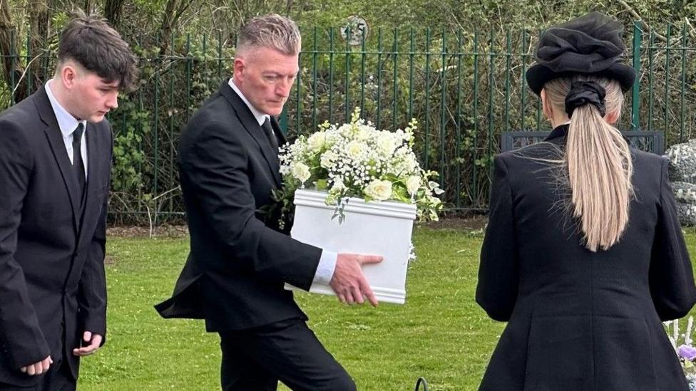 A man in a black suit carries a small white wooden coffin with a large bouquet of white flowers on its lid. Another undertake is behind him and a third, a woman in an elegant black hat, in front 
