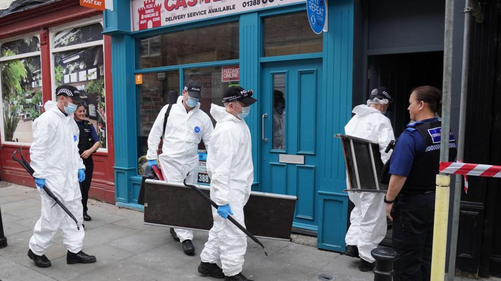 Four forensic officers in white overalls and carrying equipment enter the building where the baby's body was found in Bishop Auckland. The entrance is next to a takeaway food outlet painted in a vivid blue. It is in a row of shops. Two police staff are also in view.
