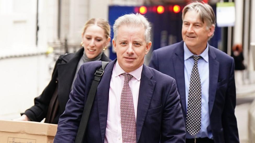 Three people in business attire walk along a narrow city street, having left court. Christopher Steele is in the foreground, wearing a dark suit with a patterned tie and carrying a shoulder bag. Behind, a woman carries a cardboard archive box with handwritten text on the side, while a man walks alongside.