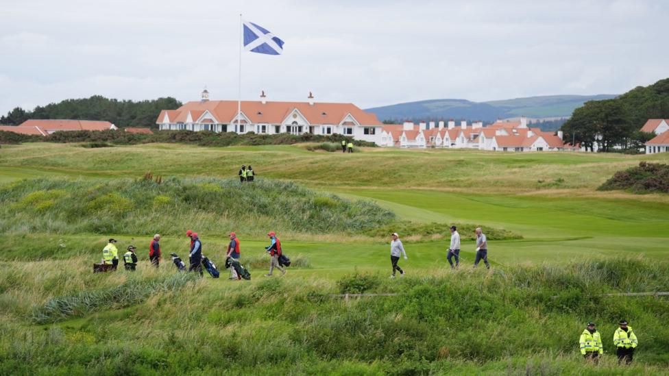 A golf course with the Scottish flag flying over the course and a large white building with a tan roof in the background. Several people are walking around the course, while several police officers stand by.
