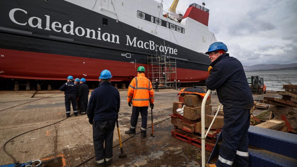 Workers make final preparations ahead of the launch of the MV Glen Rosa in April 2024. Six workers can be seen in the foreground with their back to the camera. The white, black and red ferry, with Caledonian MacBrayne written on the side, is in the distance.