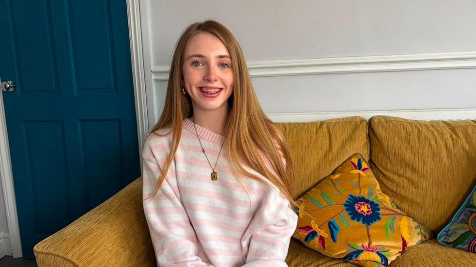 Betsi smiles at the camera. she has blonde hair and braces, she is wearing a pink and white striped top and is sat on a yellow sofa. there is a blue door in the background.