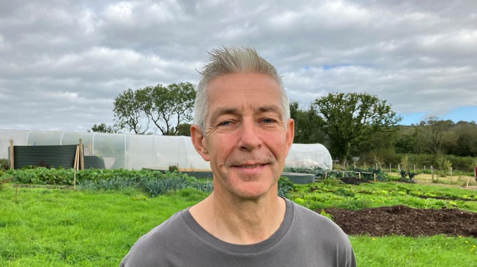 An man with grey hair and a grey, branded t-shirt standing next to a bed of vegetables