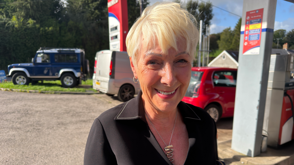 A woman with short blonde hair, wearing a black blouse, stands in a petrol station forecourt