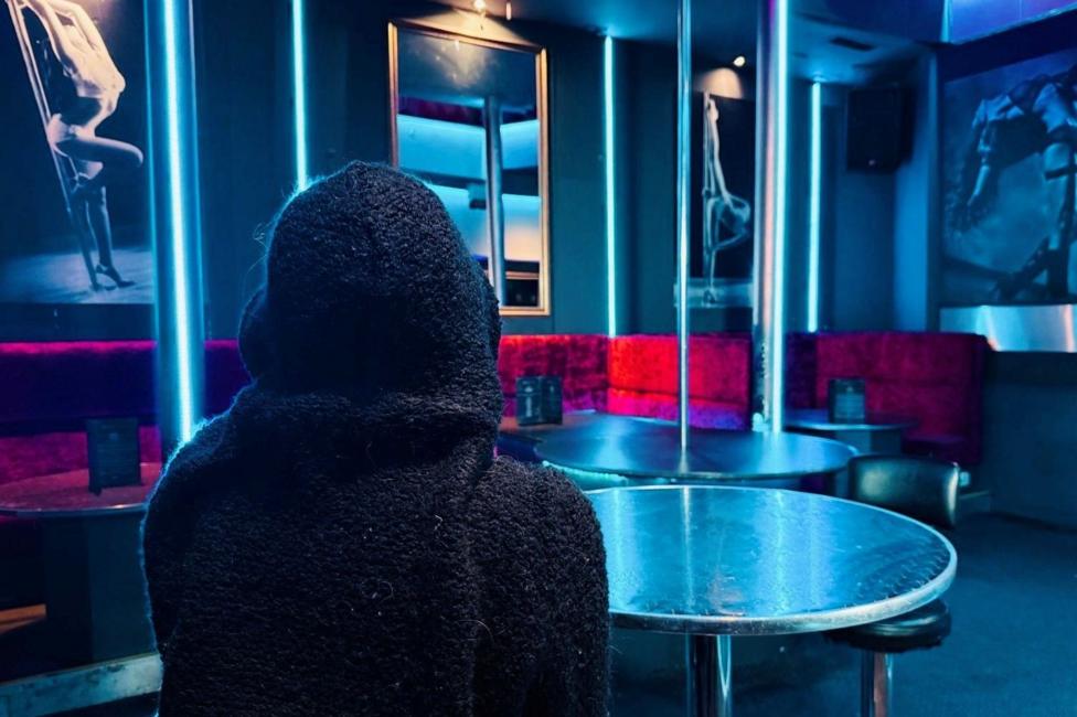 A woman photographed from behind with a hoodie covering her head, taken sitting in a strip club.