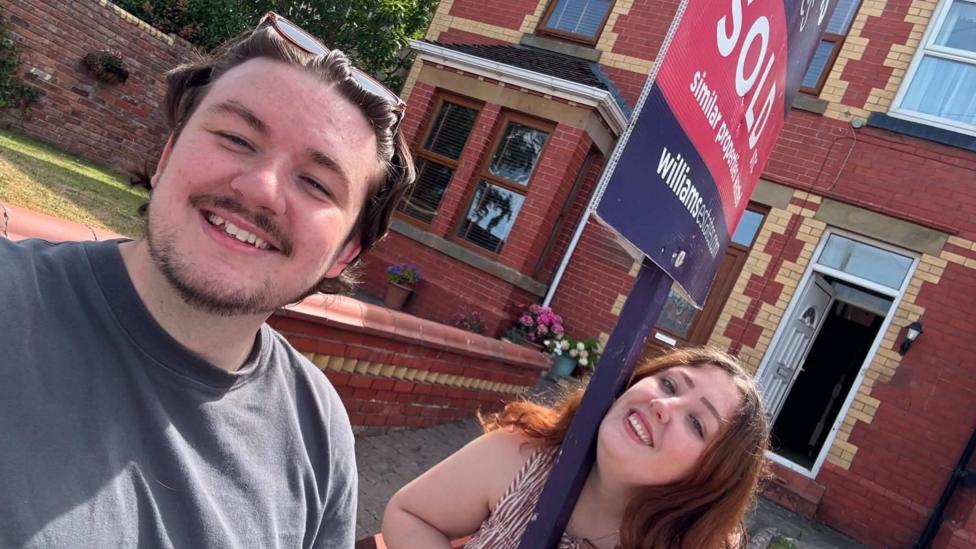 Bronya and George are standing in front of a 'sold' sign outside their red-brick terraced home. They are both smiling at the camera.