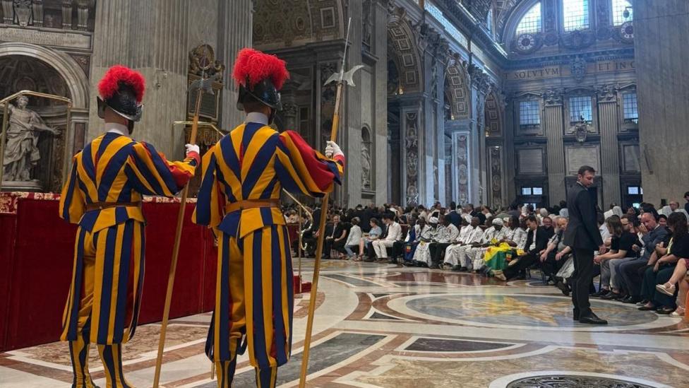 Inside St. Peter's Basilica. The picture features the Swiss Guards, they are wearing yellow and blue stripped uniforms. Behind them is a large crowd of people in front of them.