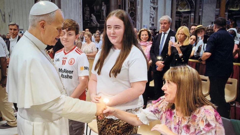 Pope Leo wearing all white is shaking hands with Eoghan and Áine's mum, Edel. She is wearing a pink flowery top and has long fair hair. The pope is looking at her and smiling.