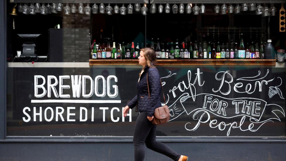 A woman walks past a Brewdog bar in London. On the glass is written Brewdog Shoreditch and Craft Beer for the People.